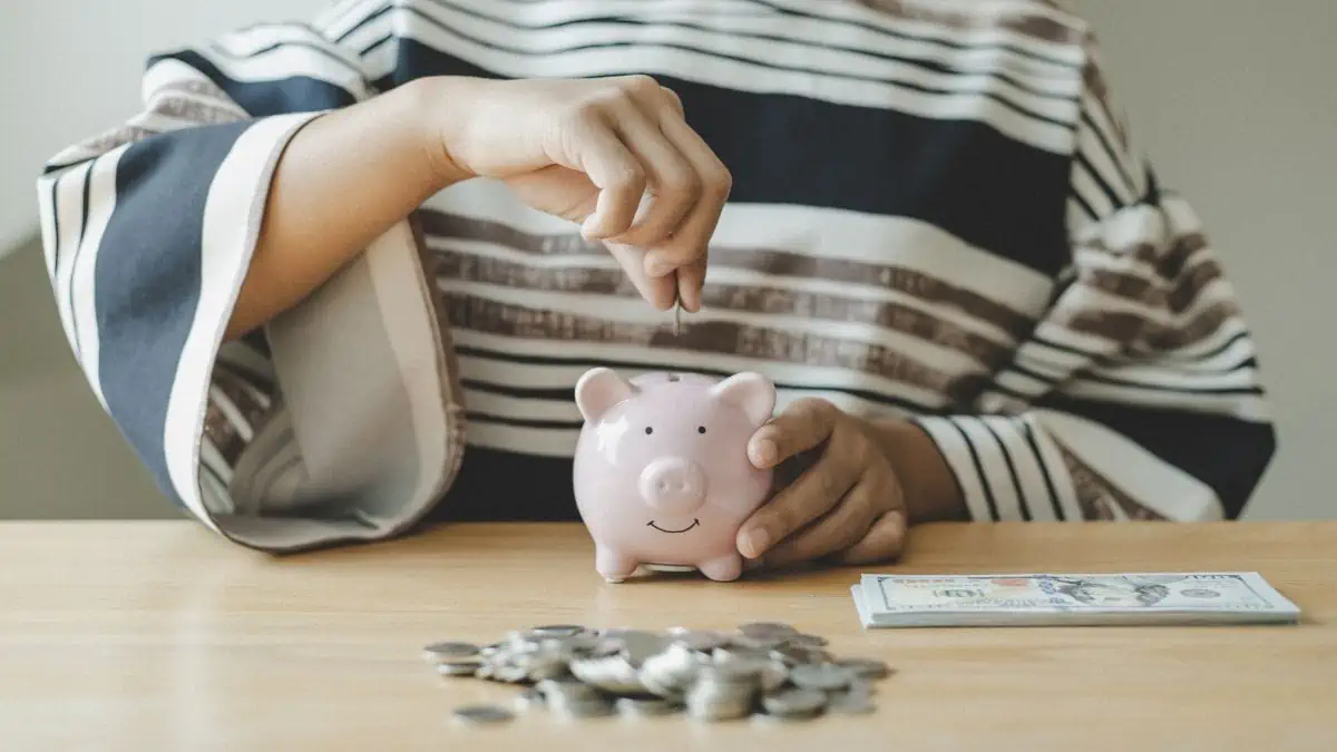 A photo of a person putting in a coin inside a piggy bank and some more coins and paper bills on the table.